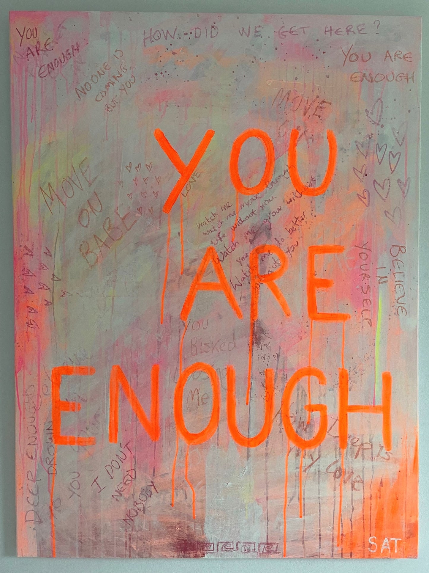 The painting 'You Are Enough' being displayed on a white wall. This piece has an abundance of words and sentances that overlap, bringing meaning. With the words You Are Enough at the forefront in neon orange writing.